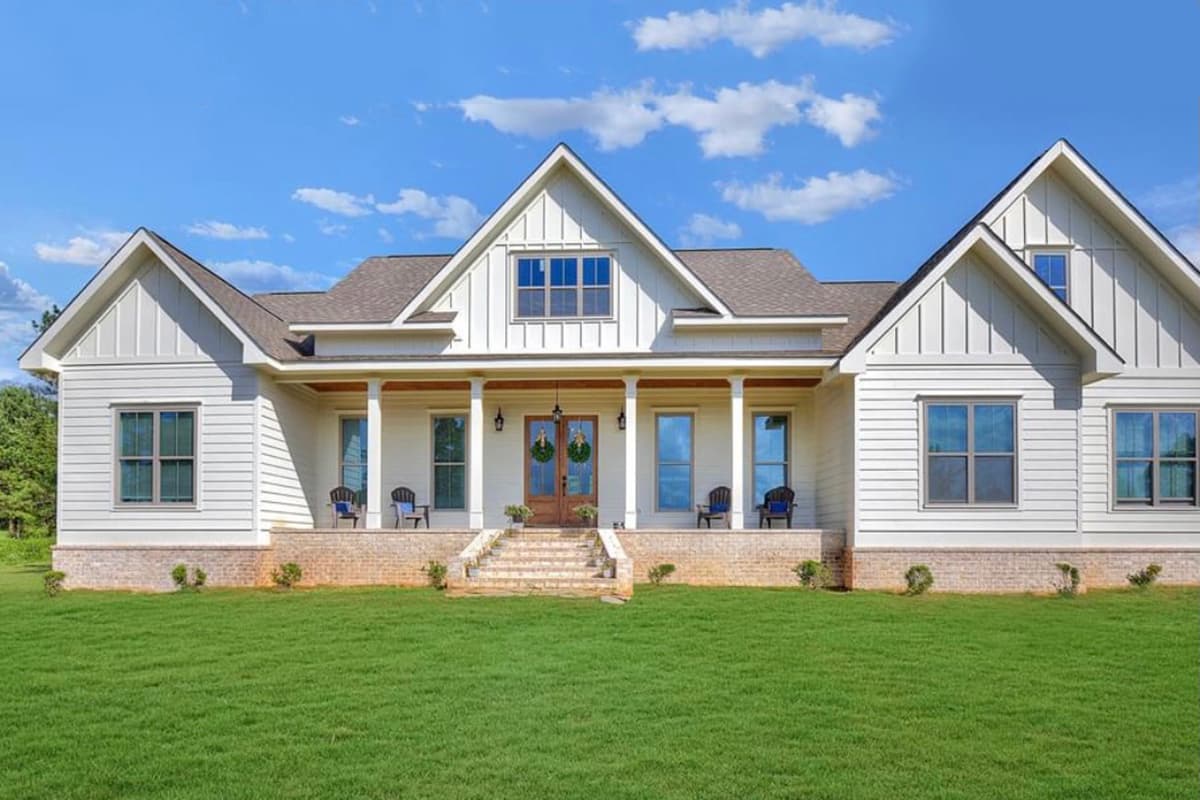 A large, white farmhouse with a wraparound porch and symmetrical design sits on a green lawn under a blue sky.