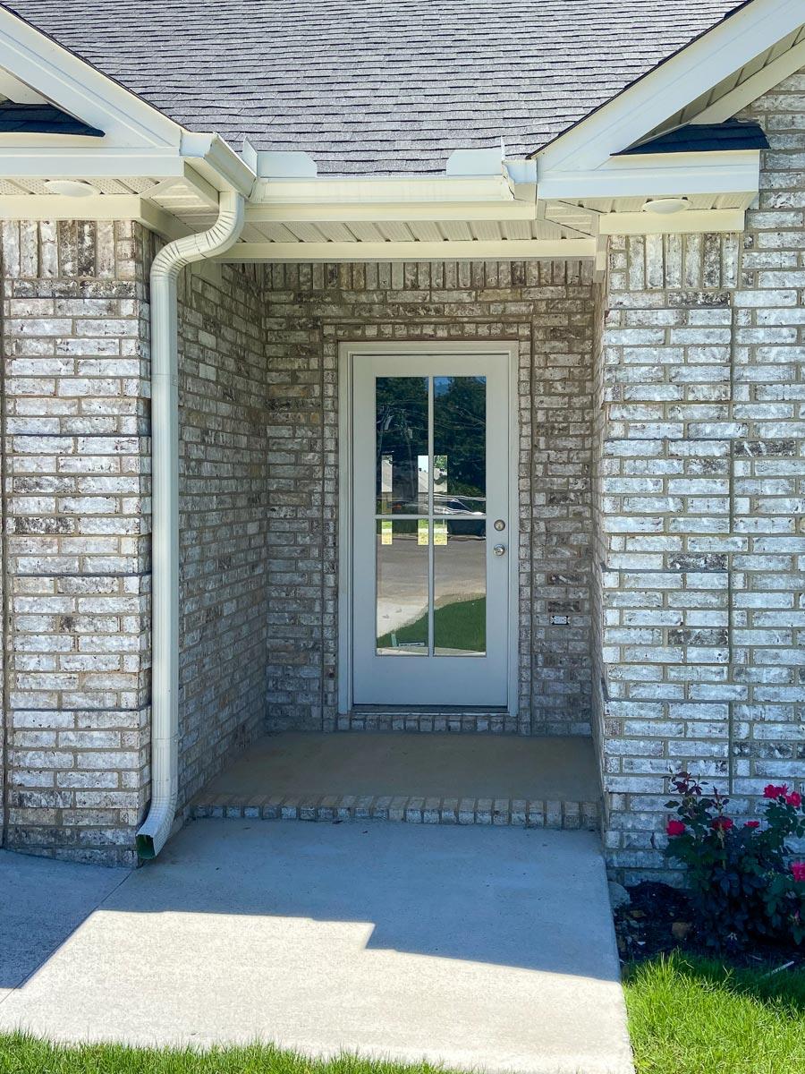 House plan exterior: White brick facade with glass-paneled door, covered entrance, and asphalt shingle roof.
