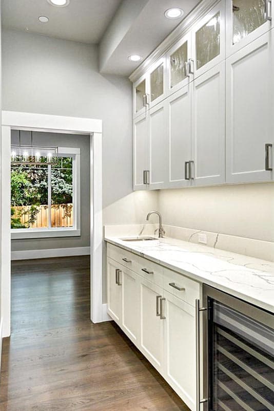 Interior view of a wet bar with white cabinets, marble countertop, sink, and wine fridge. View through doorway to backyard.