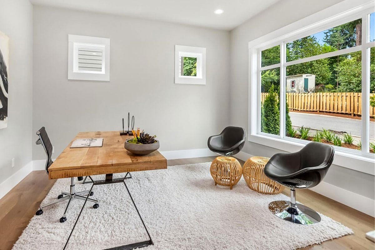 Home office with wooden desk, modern chairs, and large windows overlooking exterior. Two small square windows on wall.
