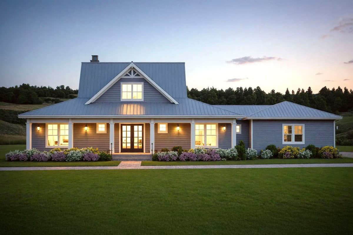 A gray house with a metal roof is lit from within, its lights contrasting with the dusk. Flower beds line the porch, in front of a green lawn.