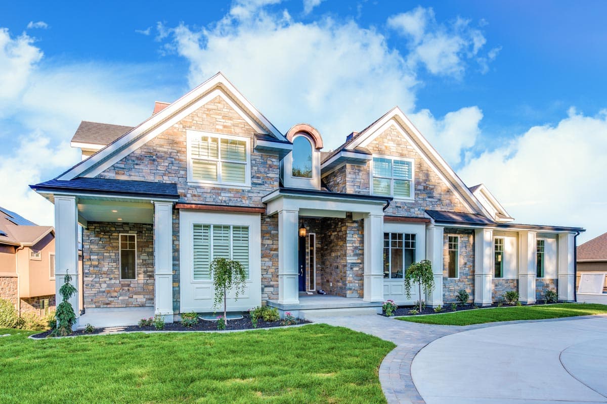 House plan exterior. Two-story home with stone facade. Features include a covered porch with columns, gabled roof, and multiple dormers.