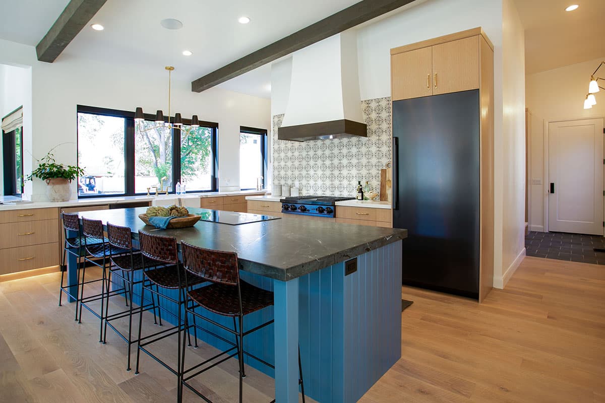 Modern kitchen with large island, blue base cabinets, dark stone countertop, decorative tile backsplash, and wood floors.