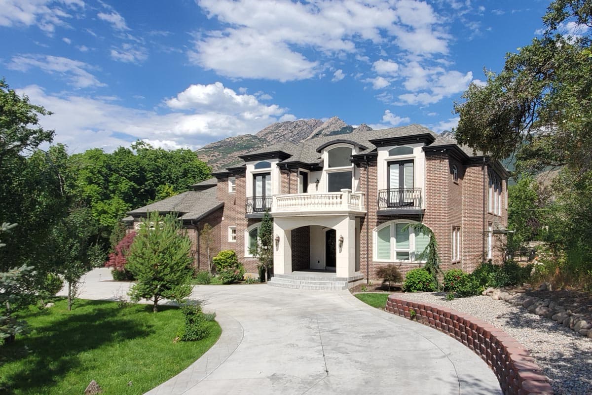 House plan exterior with brick facade, two stories, balcony, and multiple gables. Features a curved driveway and manicured landscaping.
