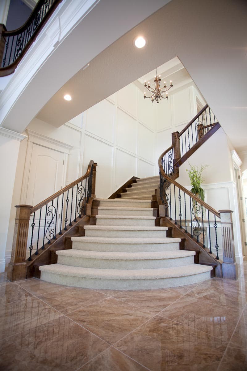 Interior view of a grand staircase with carpeted steps, dark wood banisters, ornate metal spindles, and paneled walls.