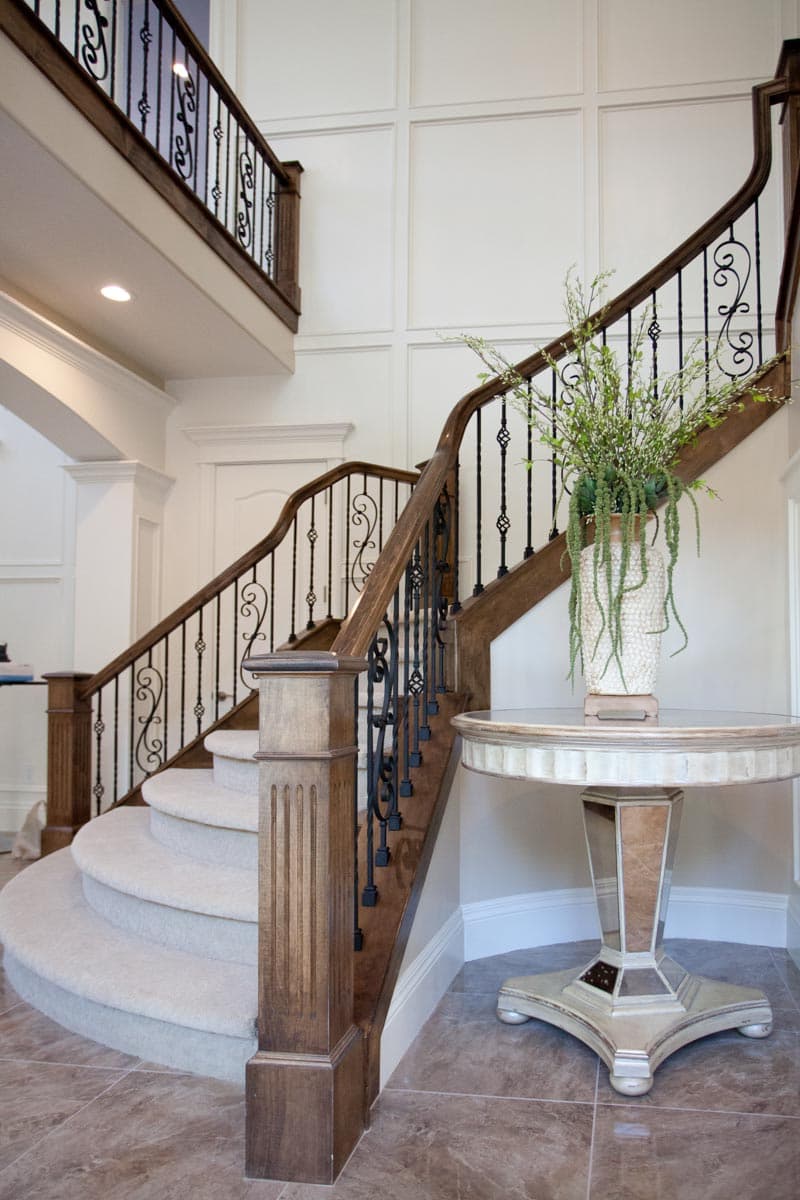 Curved staircase with wood handrails, ornate iron balusters, and carpeted steps. Decorative wall molding and pedestal table visible.