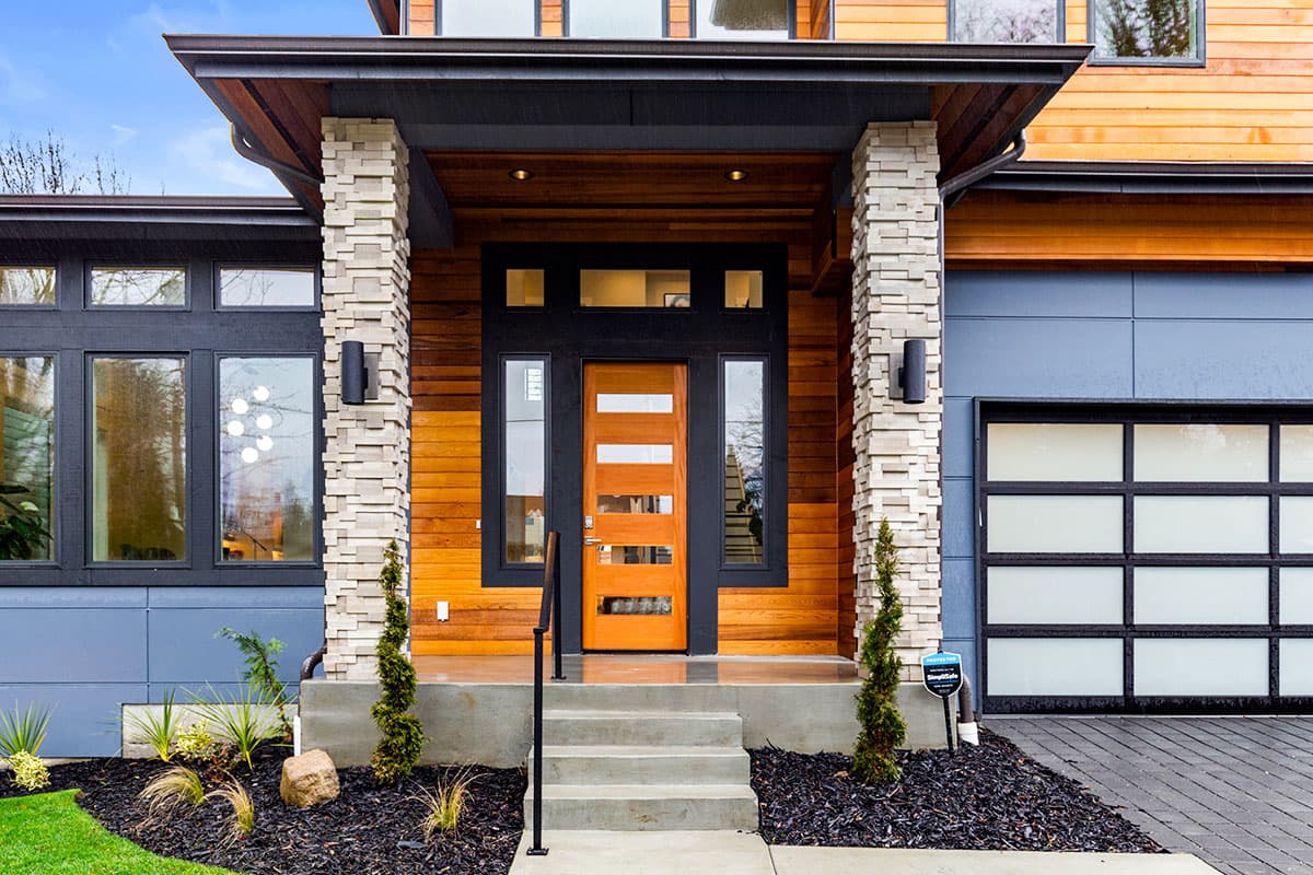 Modern house exterior with wood siding, stacked stone pillars, a glass garage door, and a covered entry with a wood door.