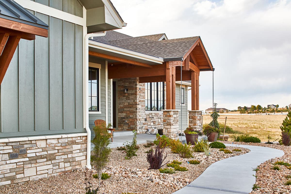 House plan exterior showing vertical siding, stone accents, covered porch with wood beams, and gabled roof.