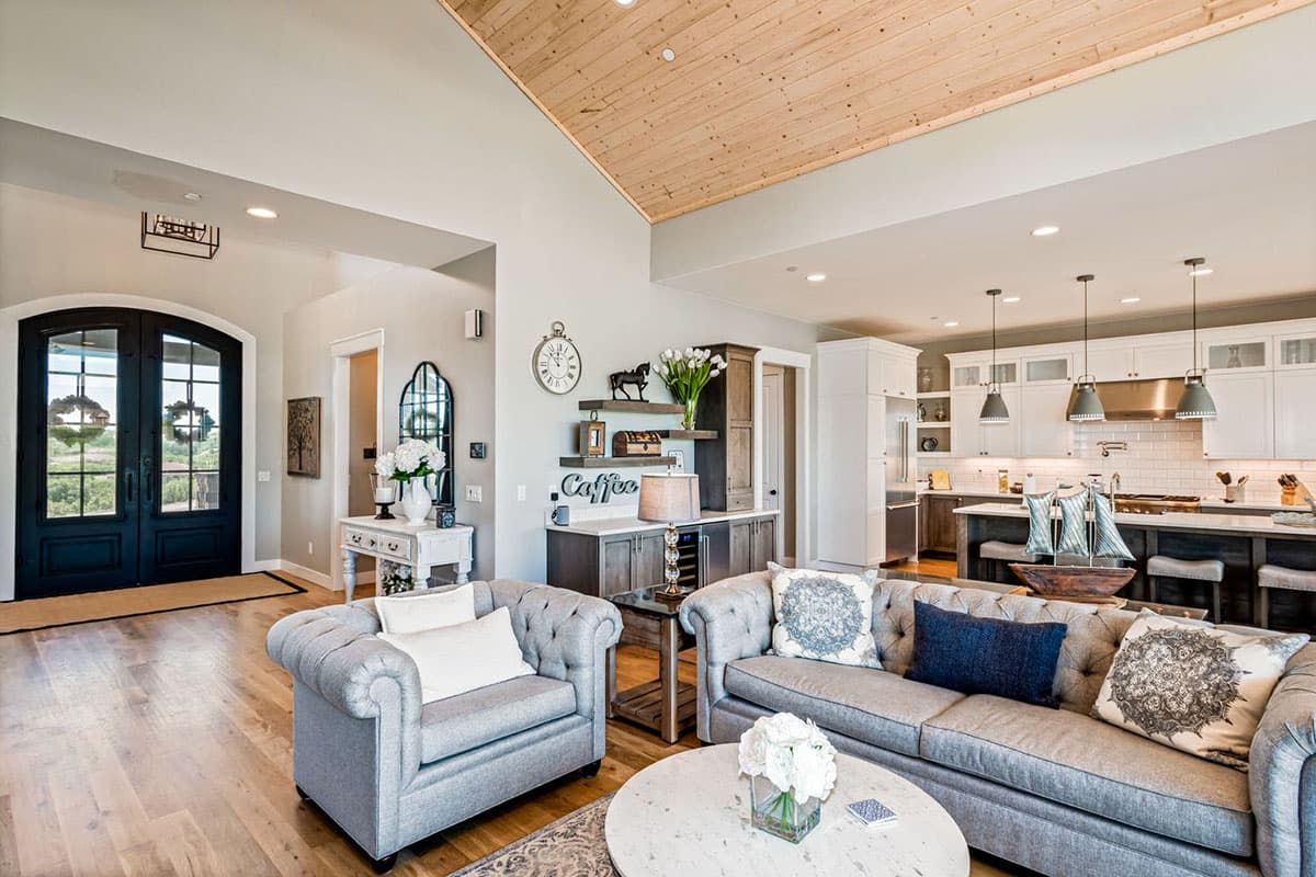 Interior view of a great room and kitchen with vaulted wood ceiling, large double doors, island, and white cabinetry.