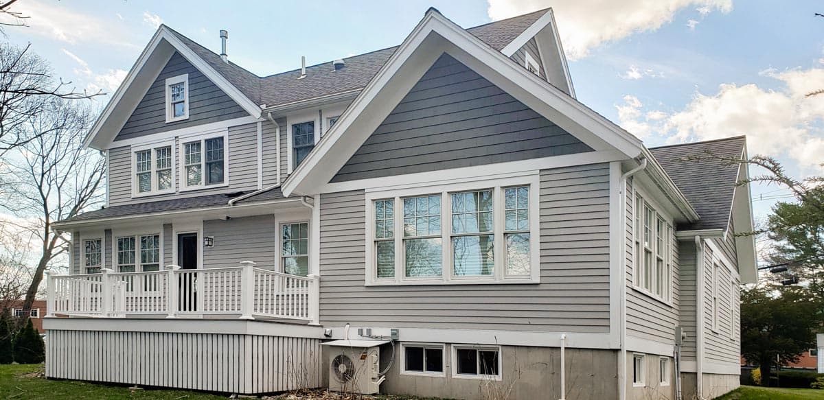 Two-story house plan exterior with gray horizontal siding, white trim, prominent gables, and a wraparound deck.