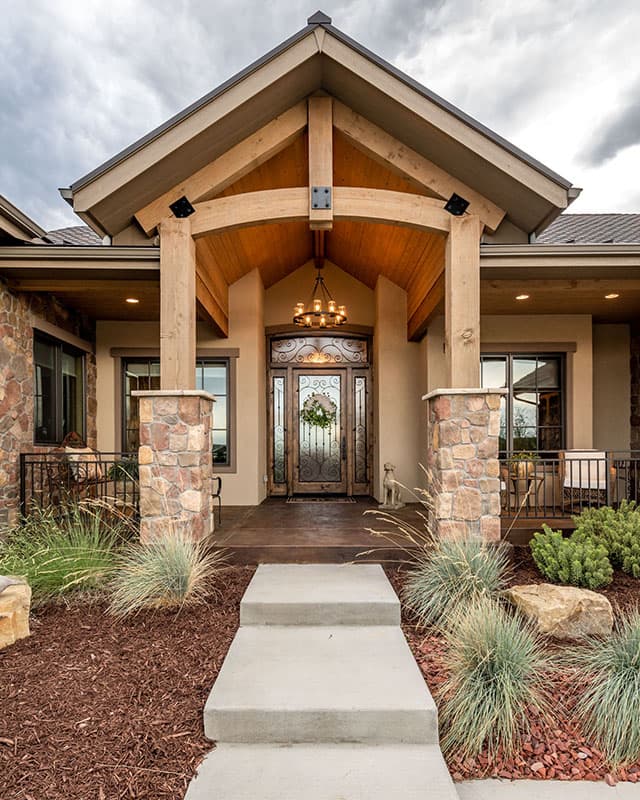 House plan exterior with prominent timber framing, stone accents, a covered porch with a vaulted wood ceiling, and a decorative metal front door.