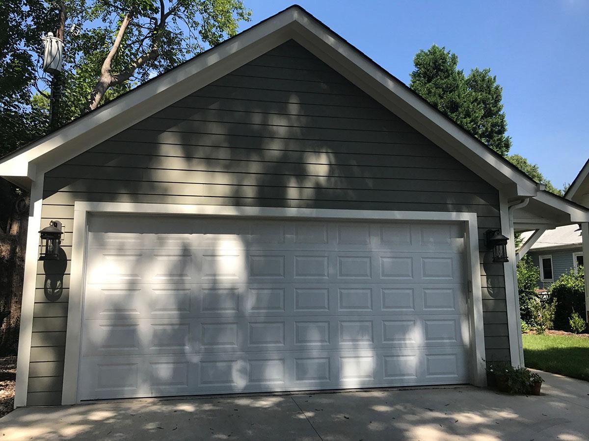 Garage exterior with white sectional door, green siding, white trim, gable roof, and two wall lanterns.