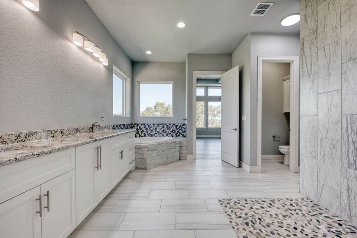 Bathroom interior with double vanity, jetted tub, and separate toilet room. Wide plank and mosaic tile flooring visible.