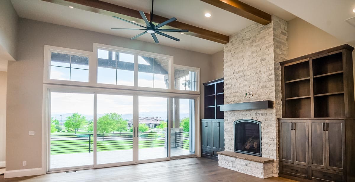 Great room with stone fireplace, built-in cabinetry, and floor-to-ceiling windows with exposed beams and ceiling fan.