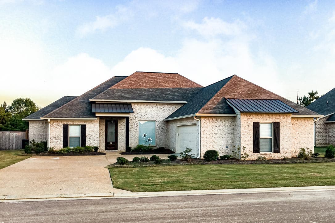 House plan exterior. Single-story, brick facade, featuring a side-entry garage, gabled roof, and decorative window shutters.