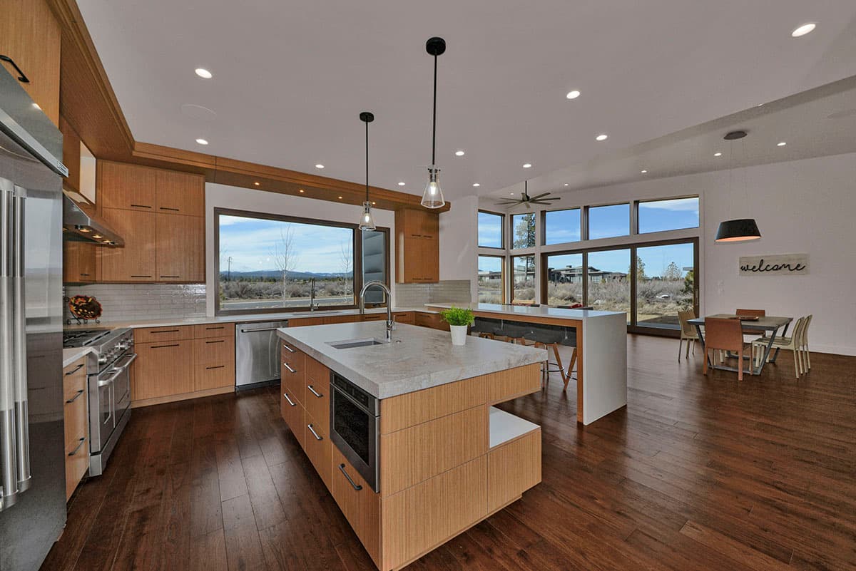 Modern kitchen with wood cabinetry, large island, stainless steel appliances, and expansive windows overlooking a scenic landscape.