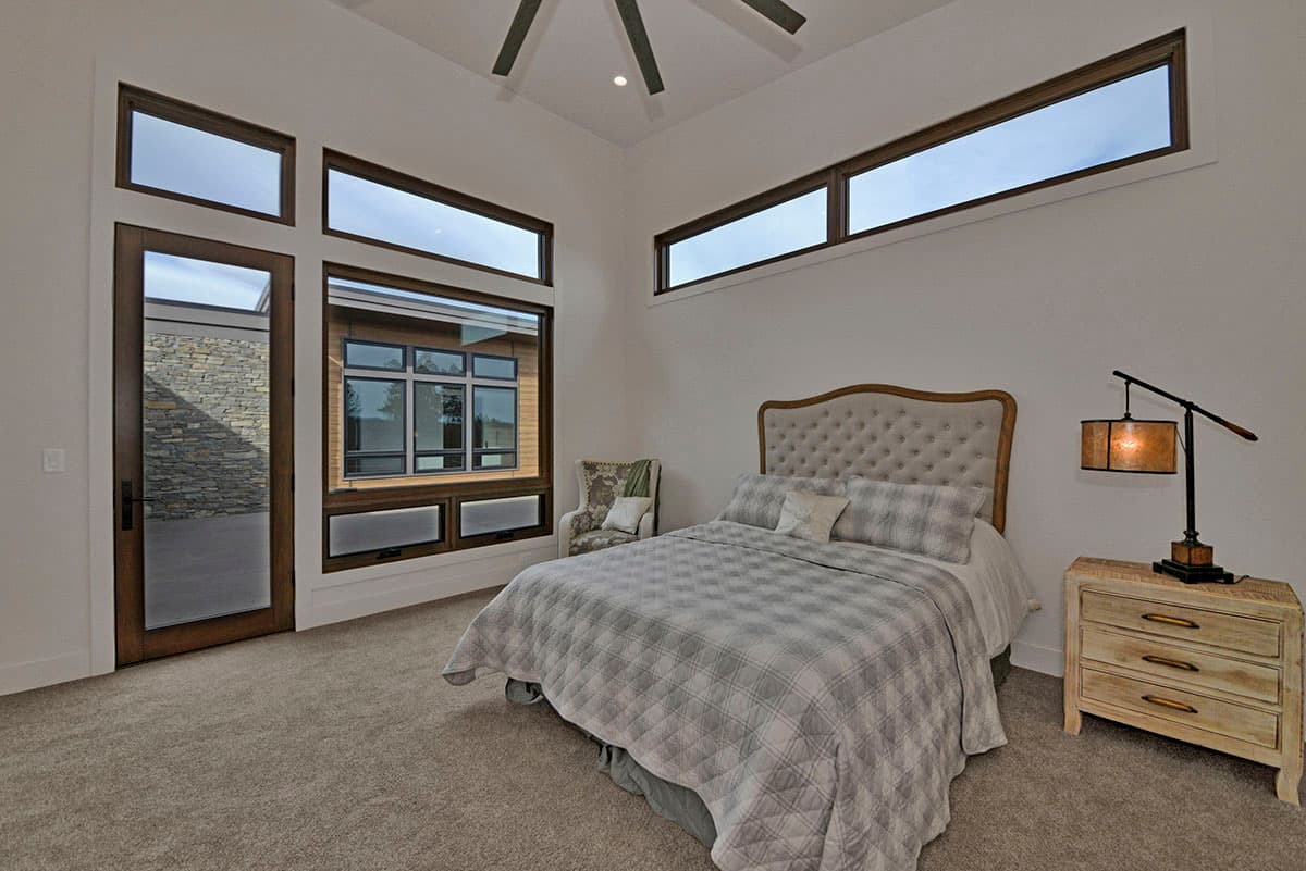 Bedroom with tufted headboard, plaid bedding, large corner windows, and glass door with stone exterior view.