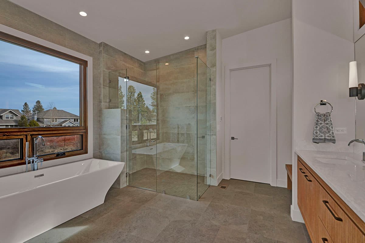 Modern bathroom with freestanding tub, glass-enclosed shower, large window, and double vanity with wood cabinets.