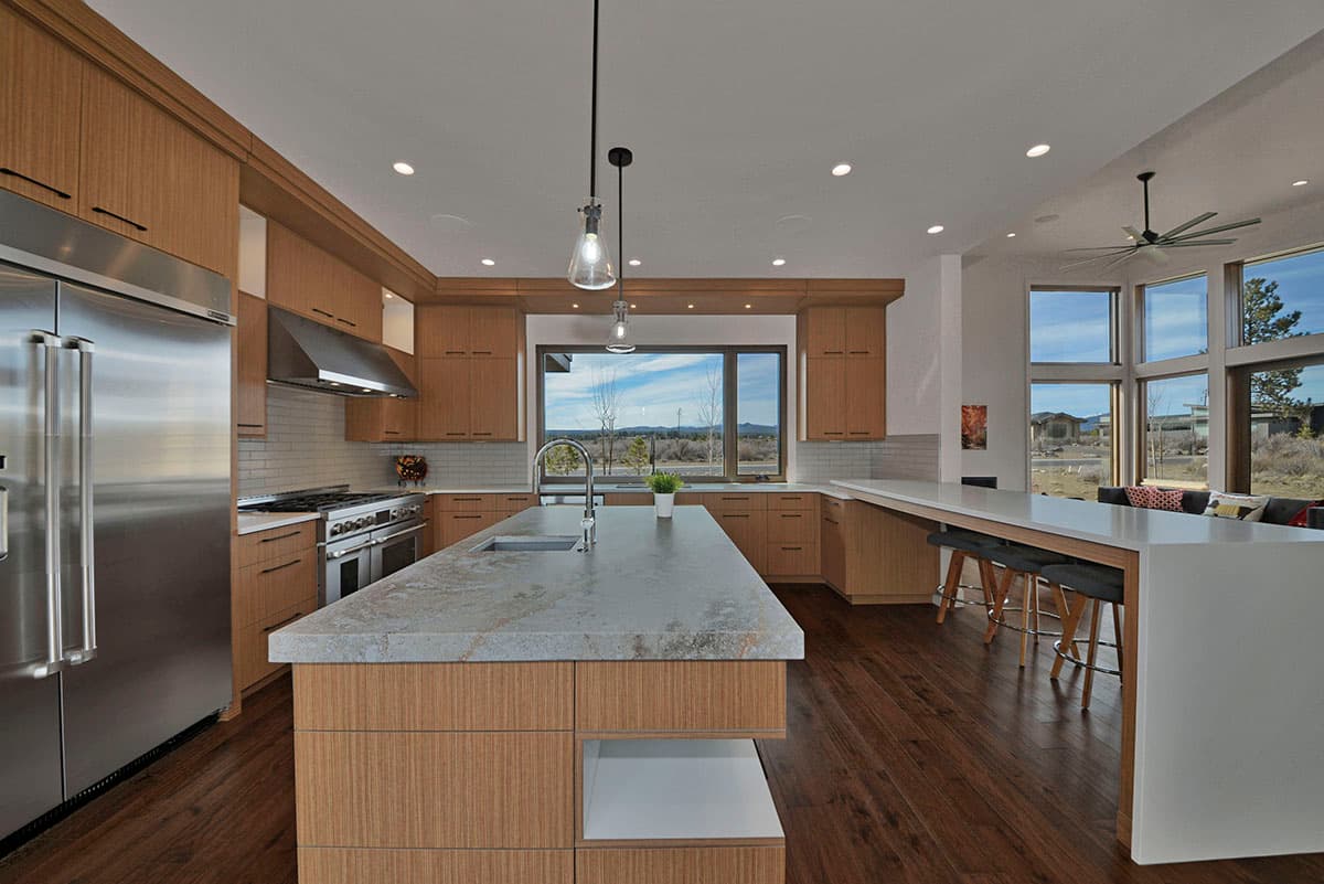 Modern kitchen interior with wood cabinetry, large island with sink, stainless steel appliances, and expansive windows overlooking the landscape.