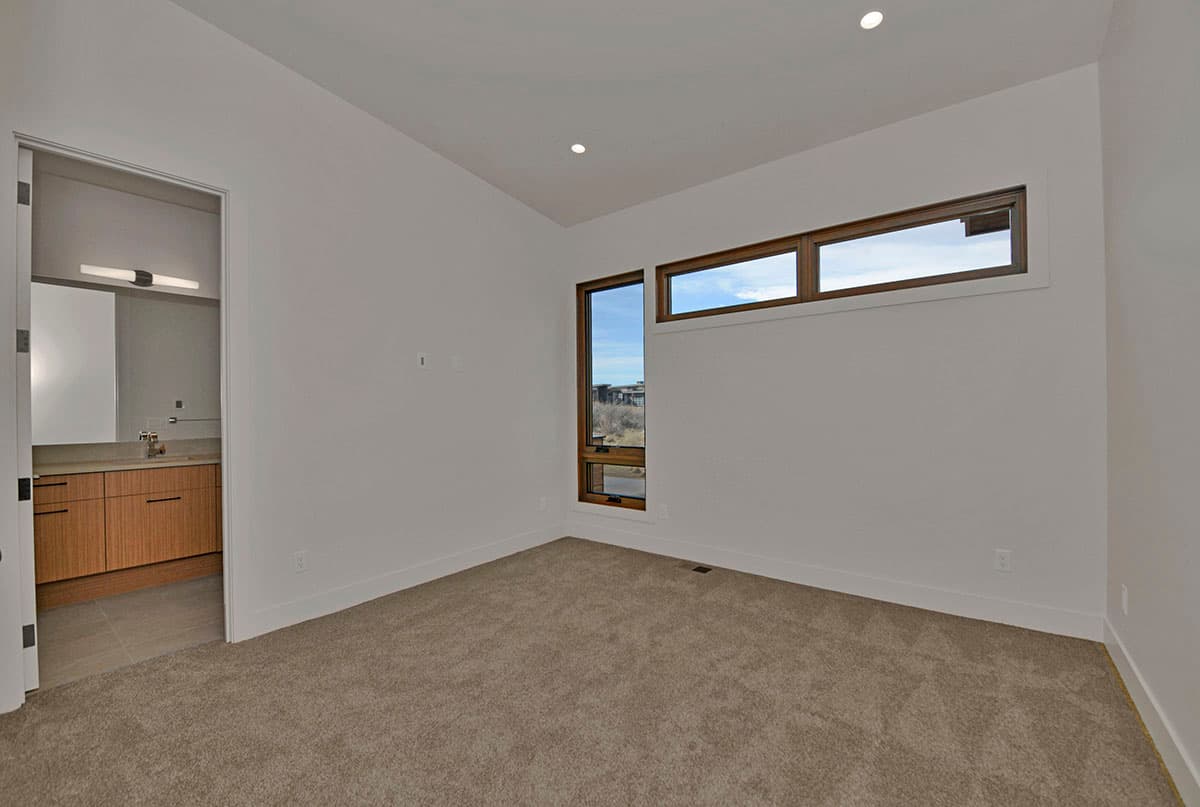 Interior view of a carpeted room with a sloped ceiling, modern windows, and an opening to a bathroom with wood vanity.