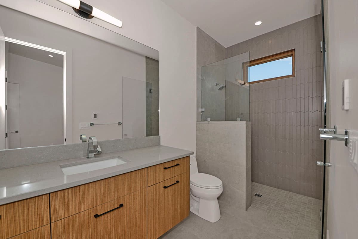 Modern bathroom with a wood vanity, granite countertop, toilet, and a glass-enclosed shower with tile accent and a horizontal window.