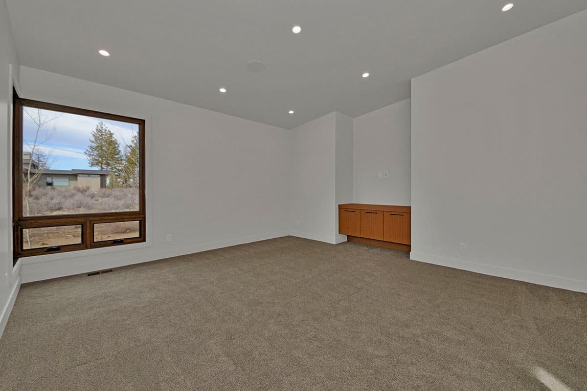Interior view of an empty room with a large corner window, carpeted floor, recessed lighting, and built-in wood cabinetry.