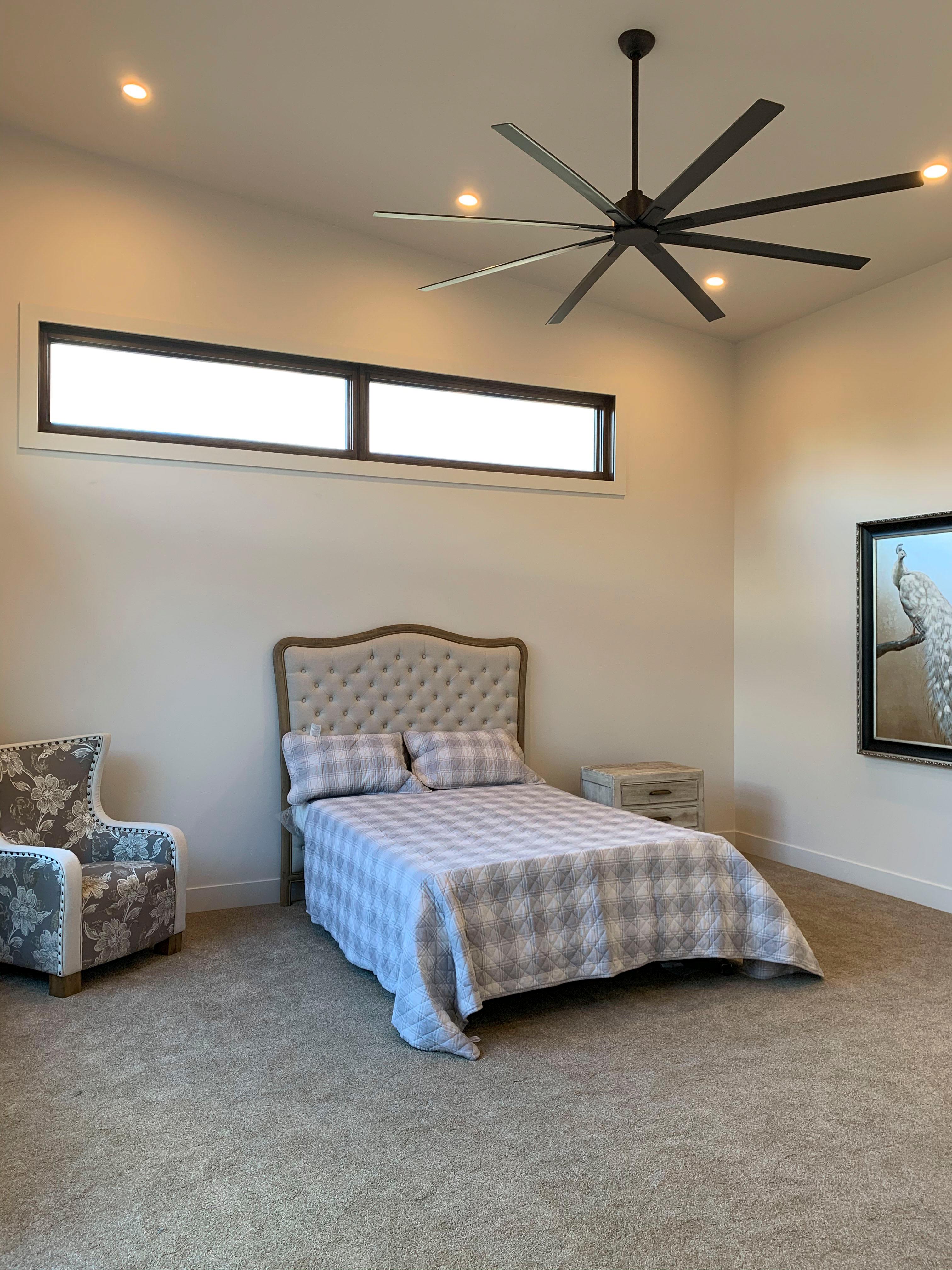 Interior rendering of bedroom with tufted headboard, patterned bedding, accent chair, large ceiling fan, and horizontal windows.