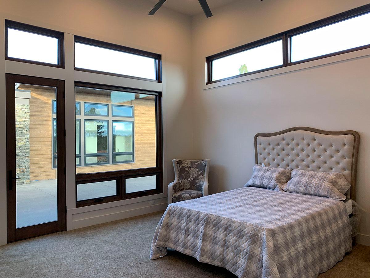 Interior view of a bedroom with a bed, tufted headboard, patterned bedding, a chair, and large windows revealing exterior wood siding and stone.