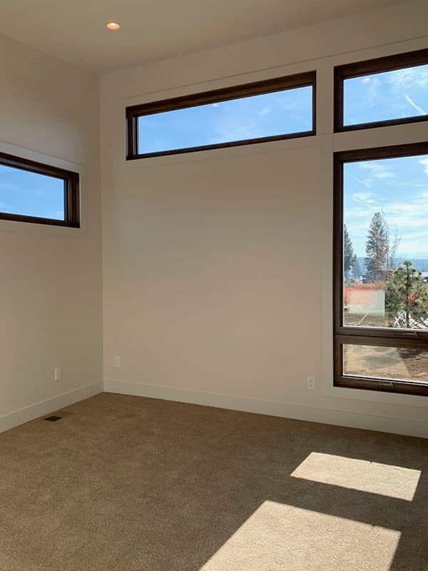 Interior view of a room with beige carpet and large, strategically placed windows with dark wood frames. Sunlight streams in.