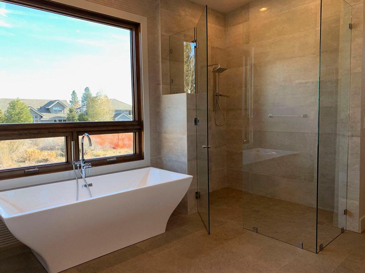 Modern bathroom with freestanding tub and glass-enclosed shower, large window with view of houses.