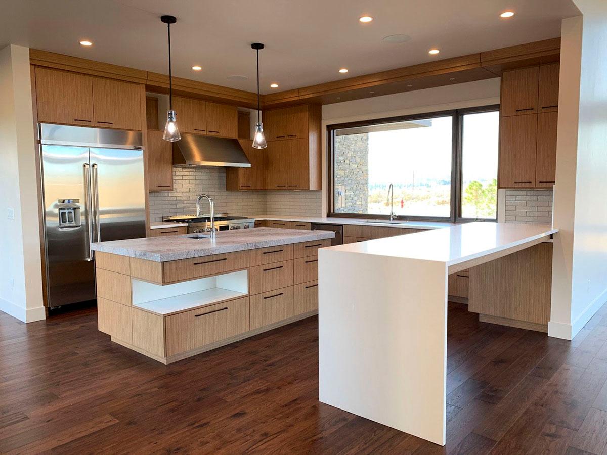 Modern kitchen with wood cabinetry, large island with waterfall edge, stainless steel appliances, and a wide window overlooking the exterior.