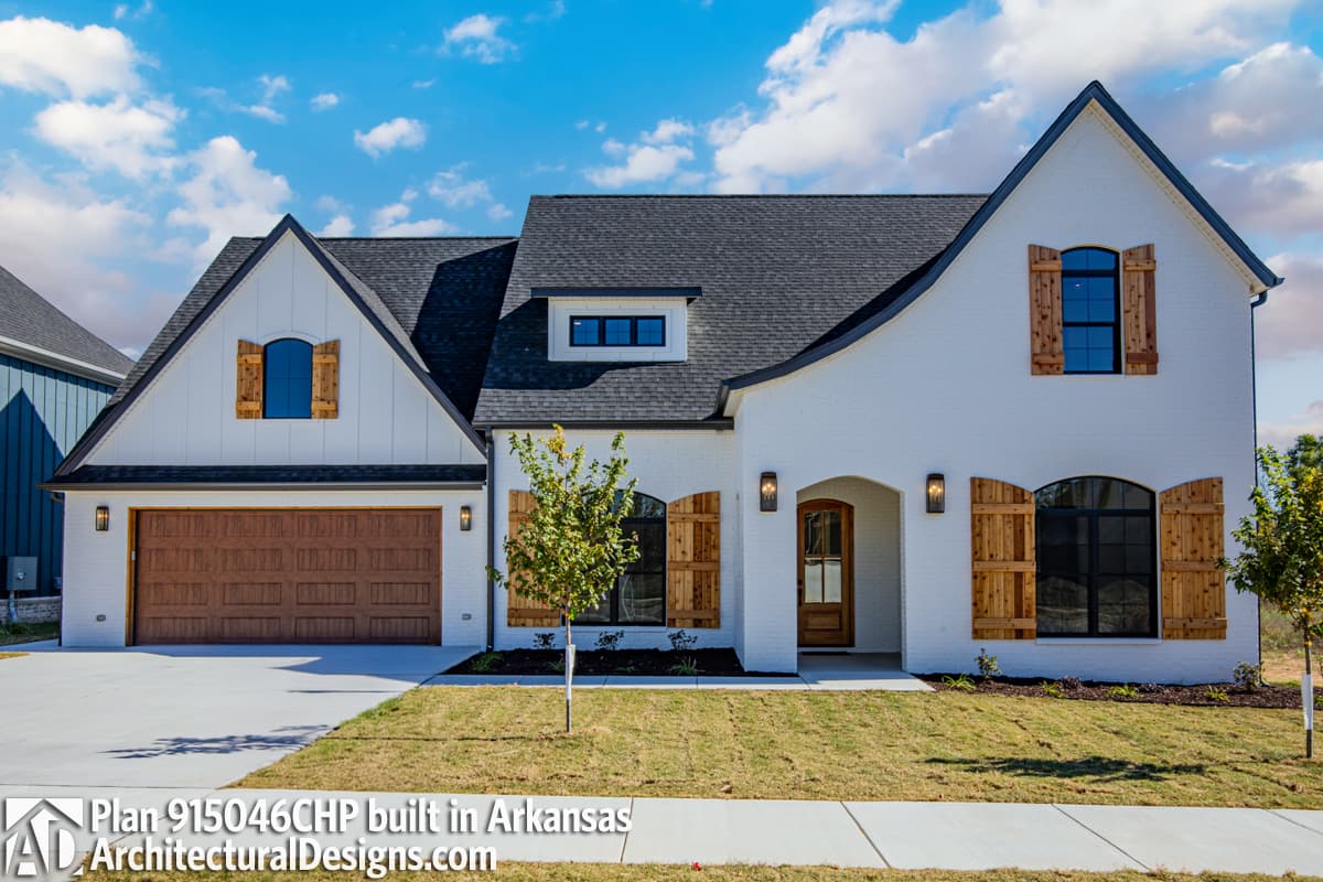 House plan exterior: Modern Farmhouse style, two stories, white brick, gabled dormer, arched windows with wooden shutters, and a side-entry garage.