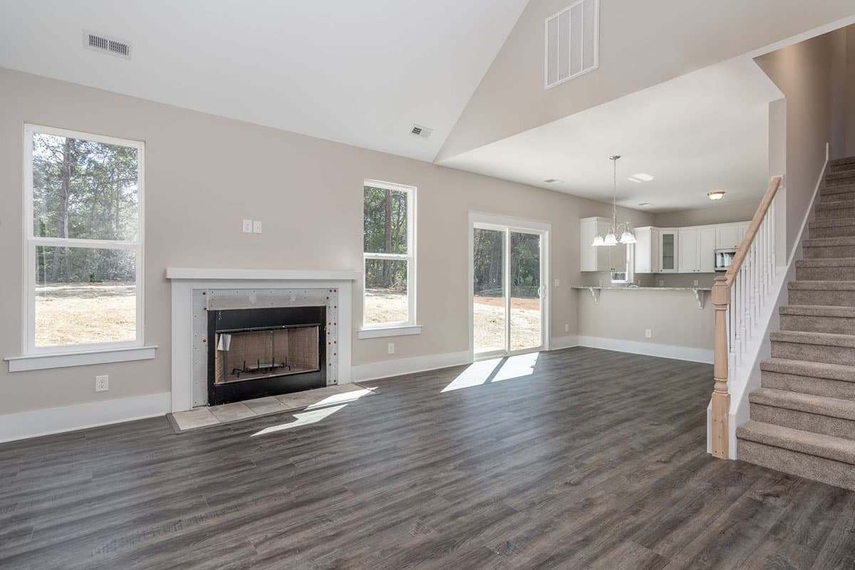 Interior view of a great room with vaulted ceiling, fireplace, staircase, and open kitchen area.