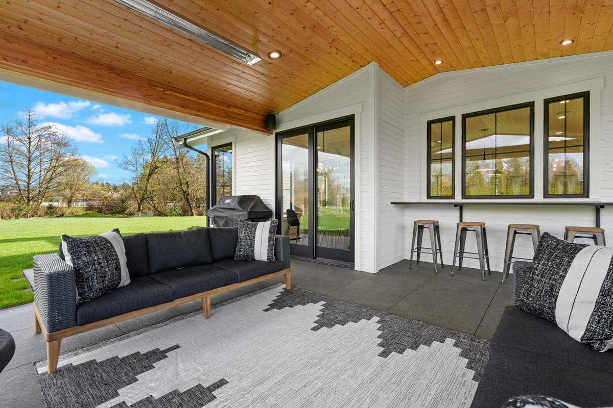 Covered patio with wood ceiling, large sliding glass doors, outdoor grill, and bar seating overlooking a lawn.