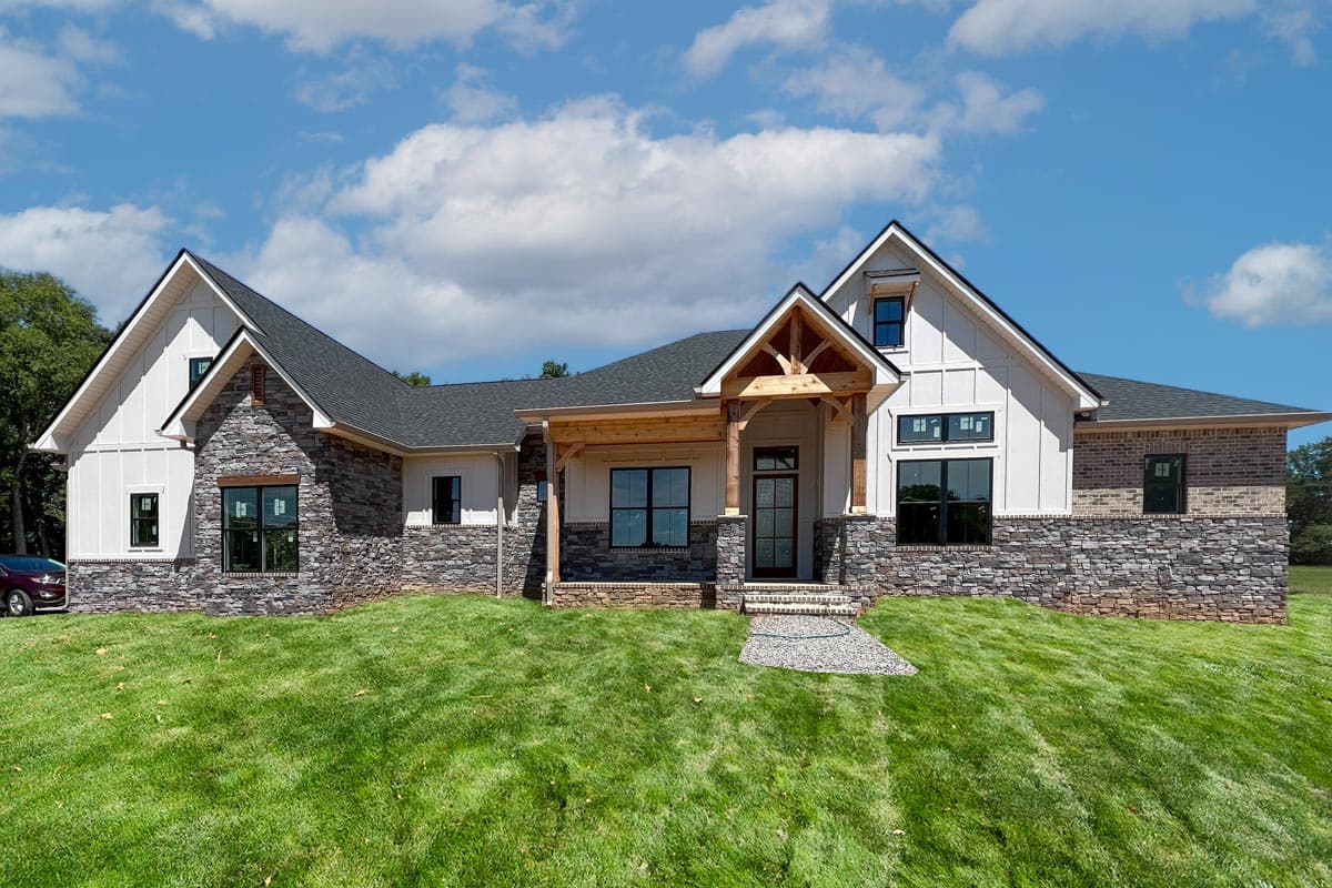 House plan exterior. Modern Farmhouse style, two stories, with a front porch, gables, stone veneer, and a dark roof. Clear, blue sky backdrop.