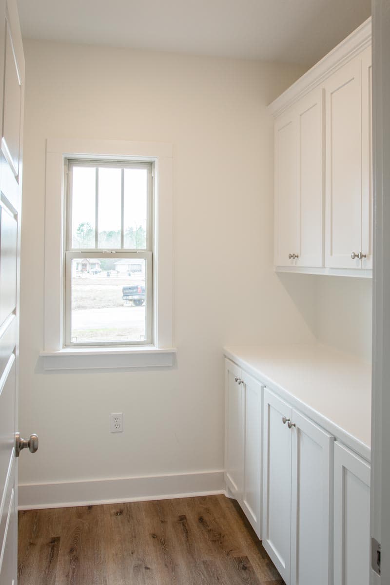Interior view of laundry room with white cabinets, countertops, and a window above wood-look flooring.