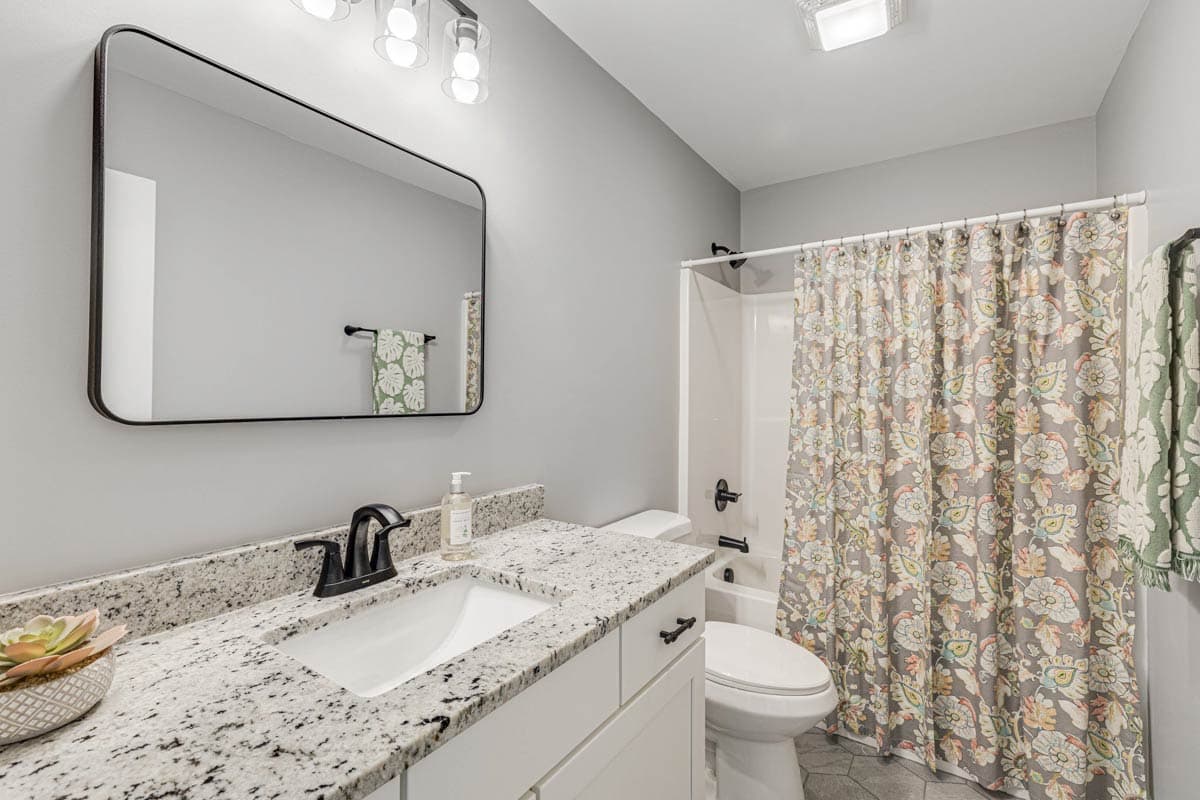 Bathroom with vanity, granite countertop, toilet, and shower with floral curtain.