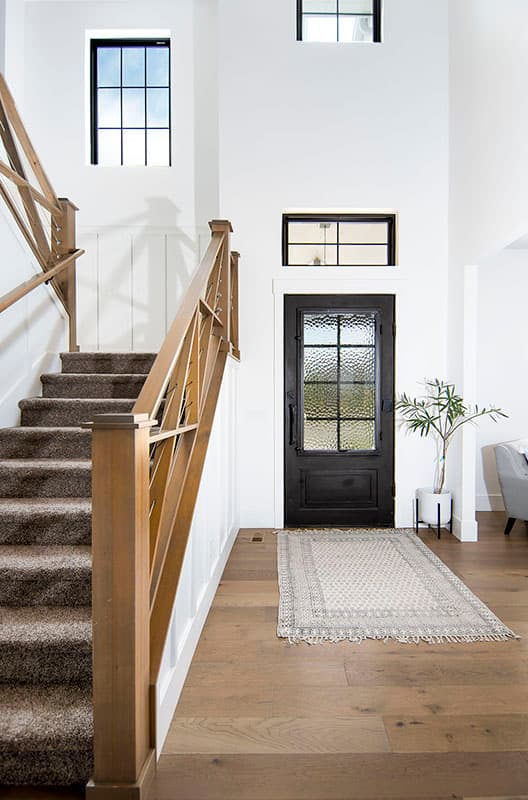 Interior view of modern entryway with carpeted staircase, black metal door with textured glass, and tall windows.