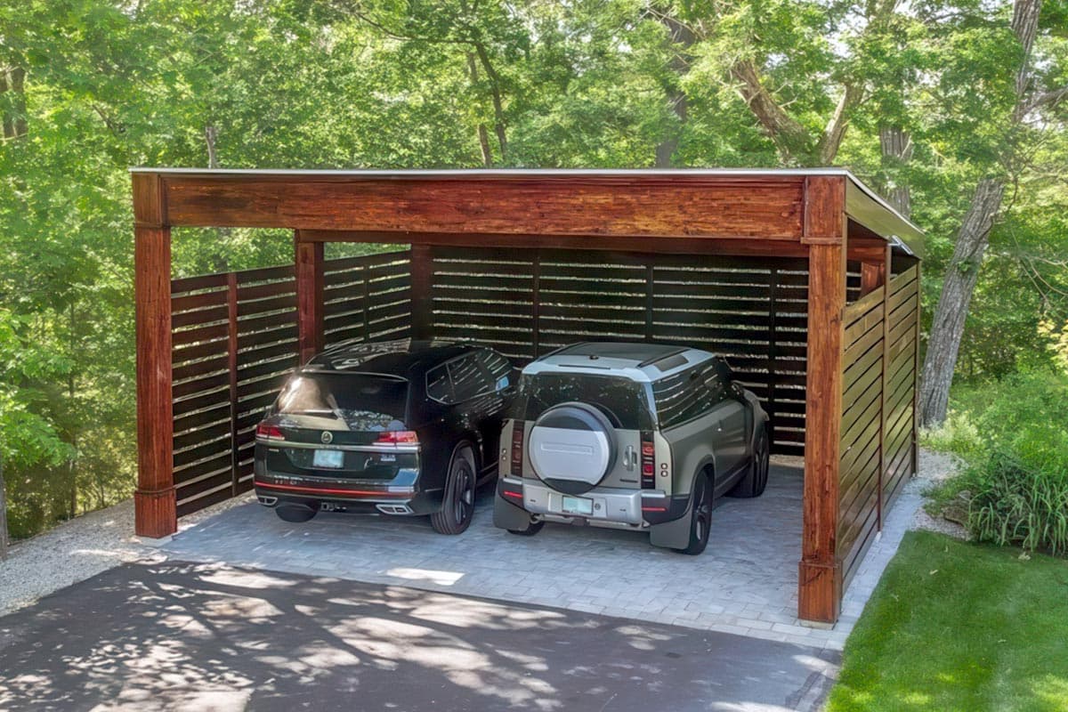 Modern carport with horizontal wood slat walls, exposed timber framing, and a metal roof. Two SUVs parked inside.