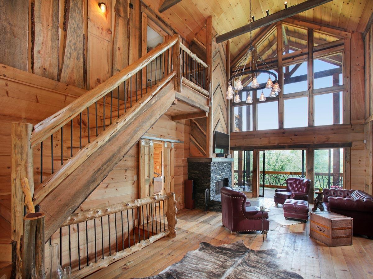 Interior living room with wood-paneled walls, a stone fireplace, a wooden staircase with metal railings, and a wall of large windows.