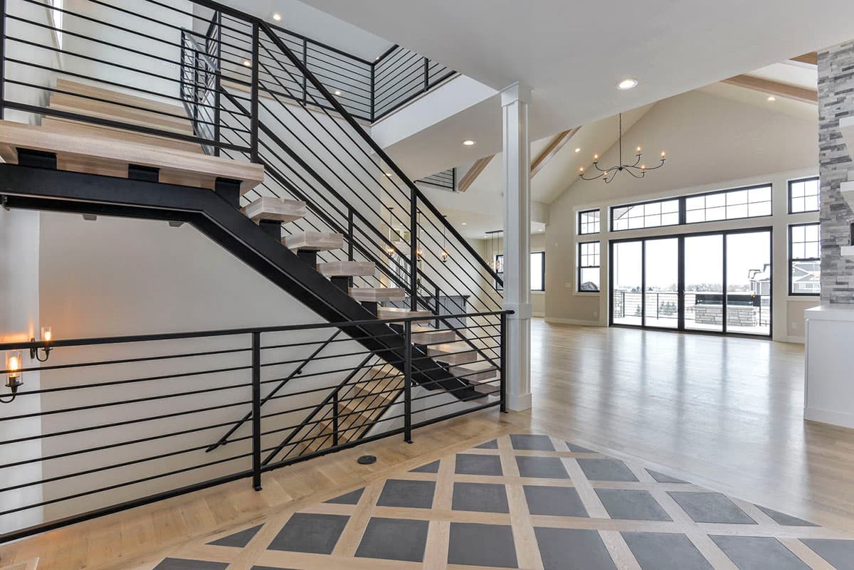 Modern house interior featuring a floating staircase with black metal railing, vaulted ceiling with wood beams, and large windows overlooking a patio.