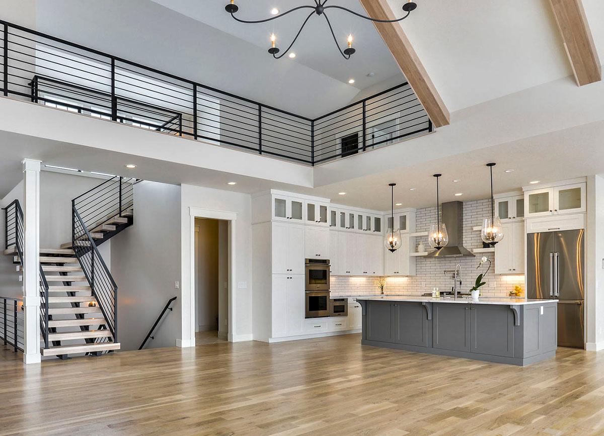 Modern open-concept kitchen with island, white cabinetry, and stainless steel appliances. Floating staircase with black railing leads to loft.