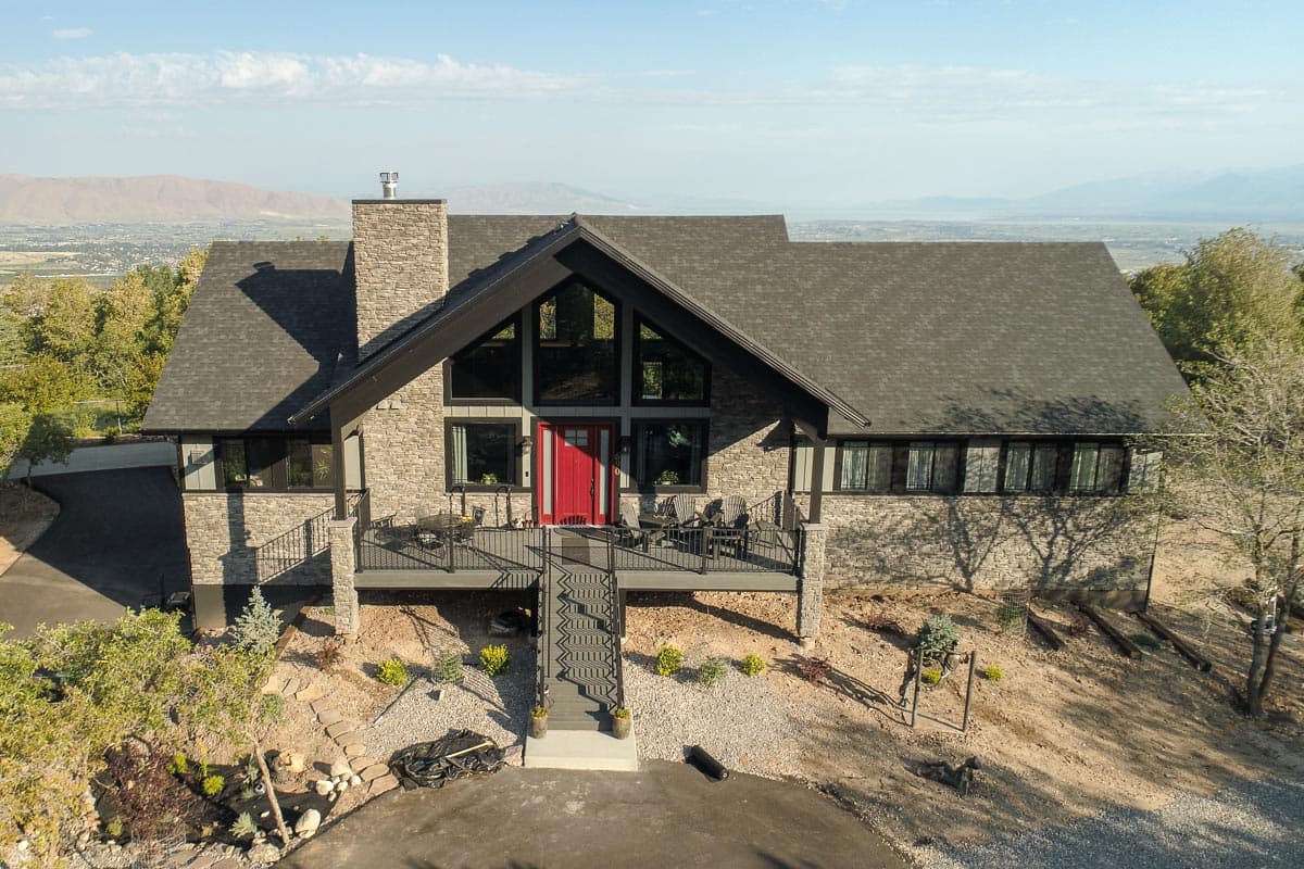 House plan exterior. Two-story home with stone facade, multi-gable roof, and a front-facing deck with access stairs leading to a red front door.