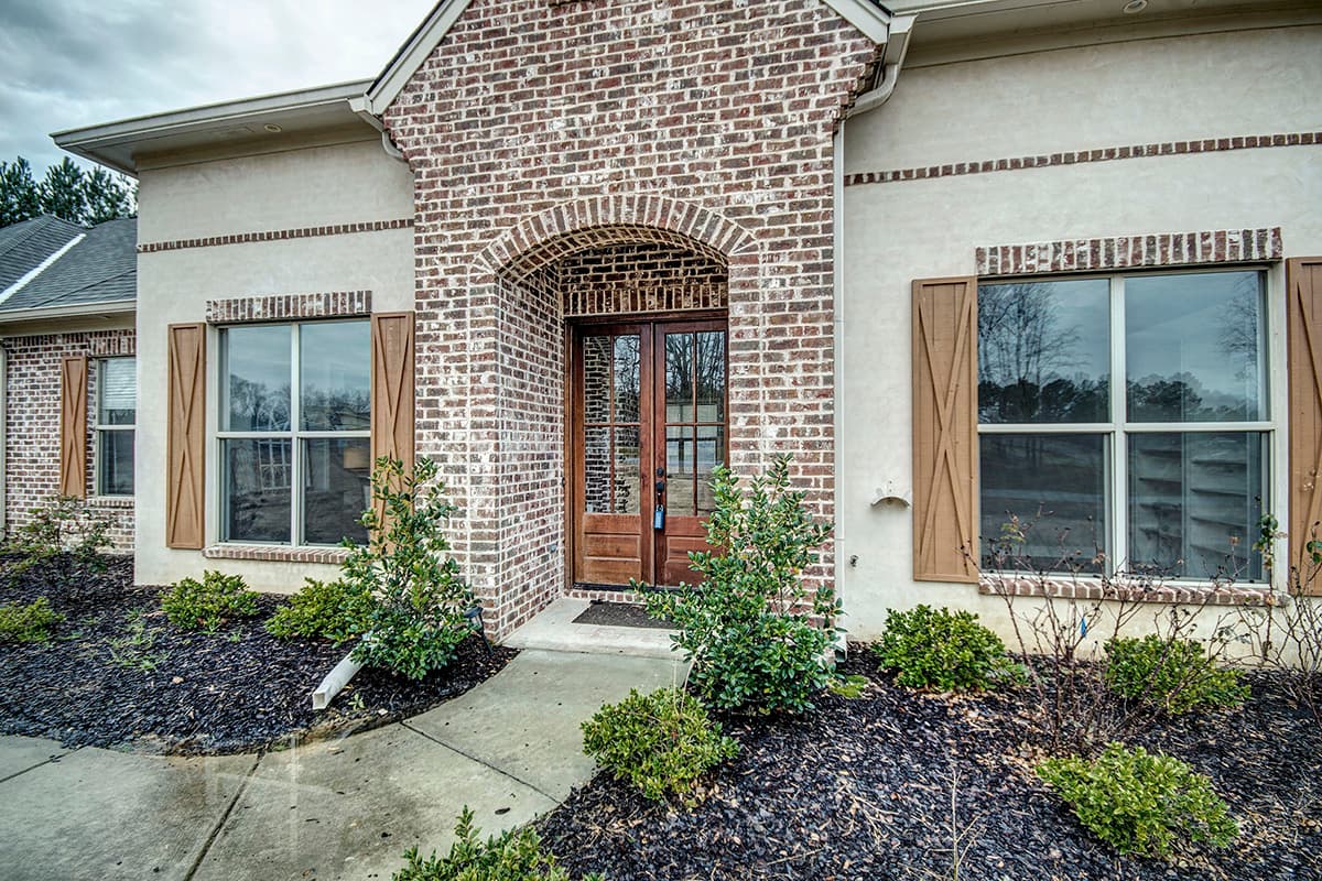 House plan exterior featuring a brick facade, arched double-door entry, and wood shutters on windows.