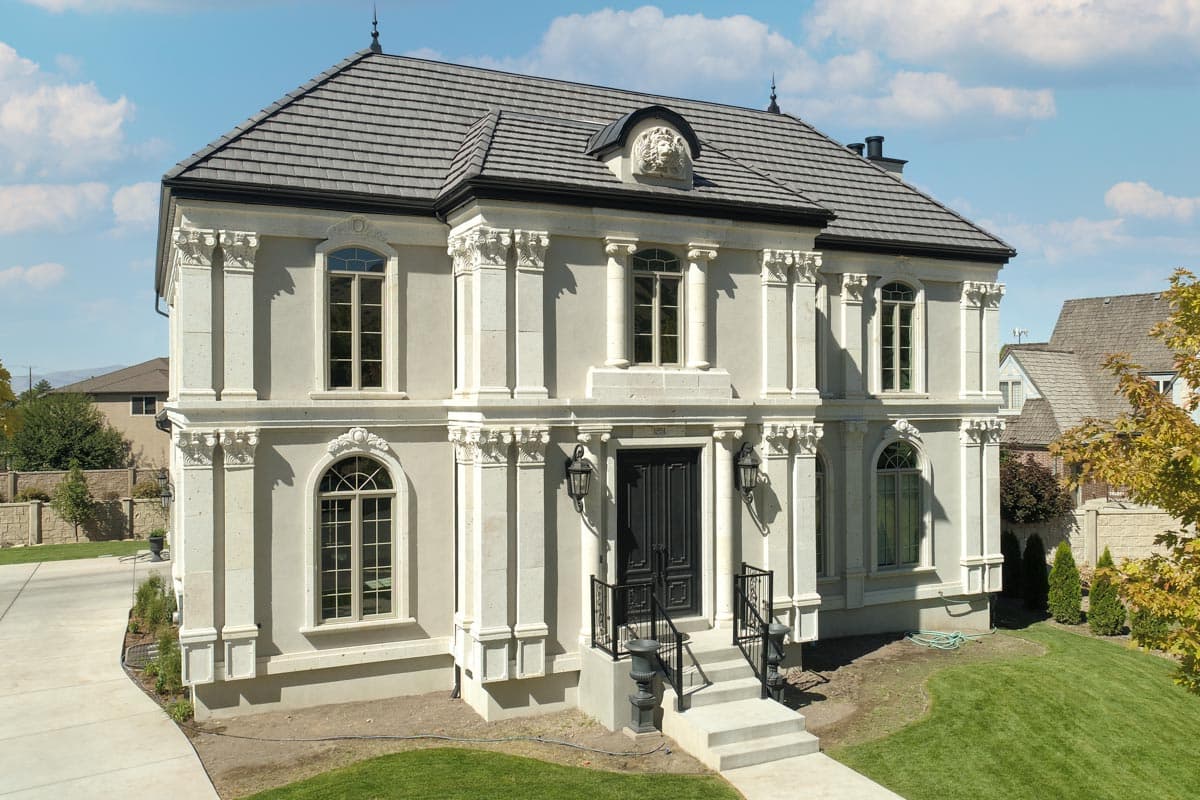 House plan exterior: Two-story, symmetrical facade with decorative columns, arched windows, mansard roof, and central dormer with sculpted relief.