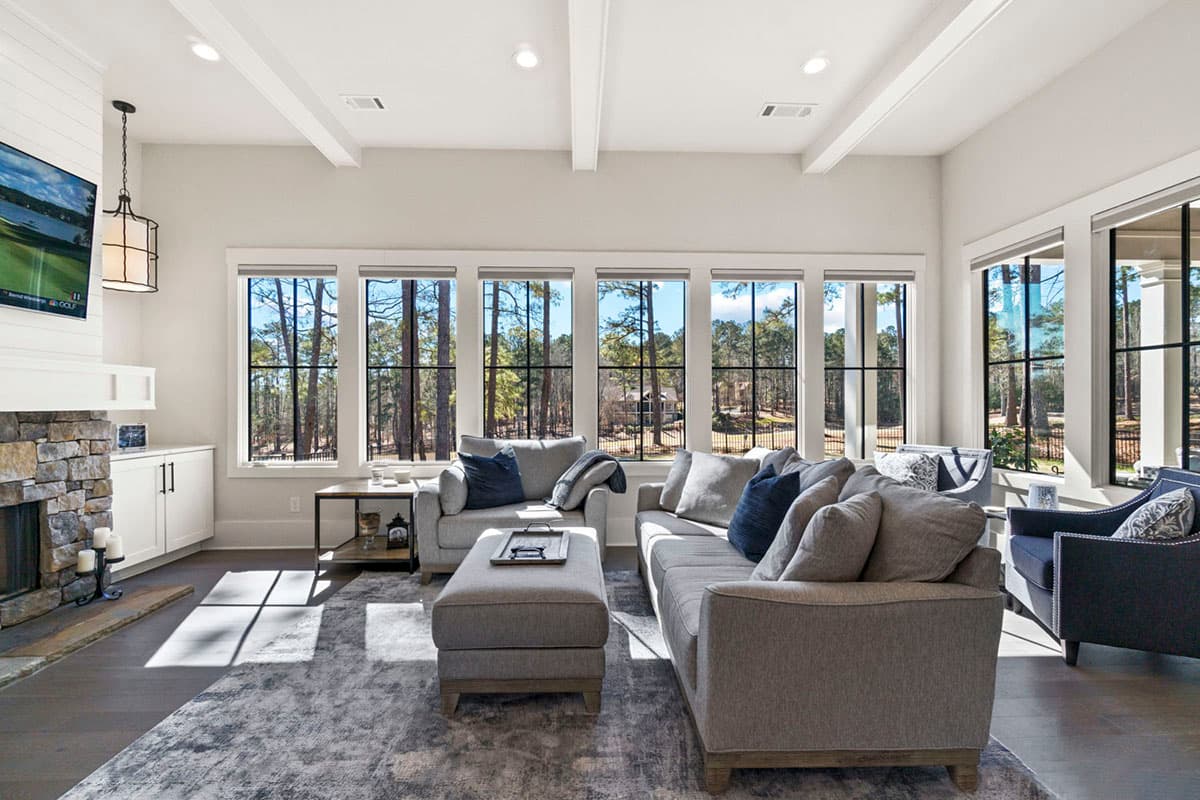 Living room with stone fireplace, vaulted beamed ceiling, and a wall of windows overlooking wooded area.