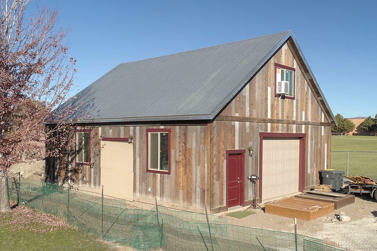 House plan exterior with corrugated metal roof, wooden siding, one garage door, two entry doors, and a dormer window.
