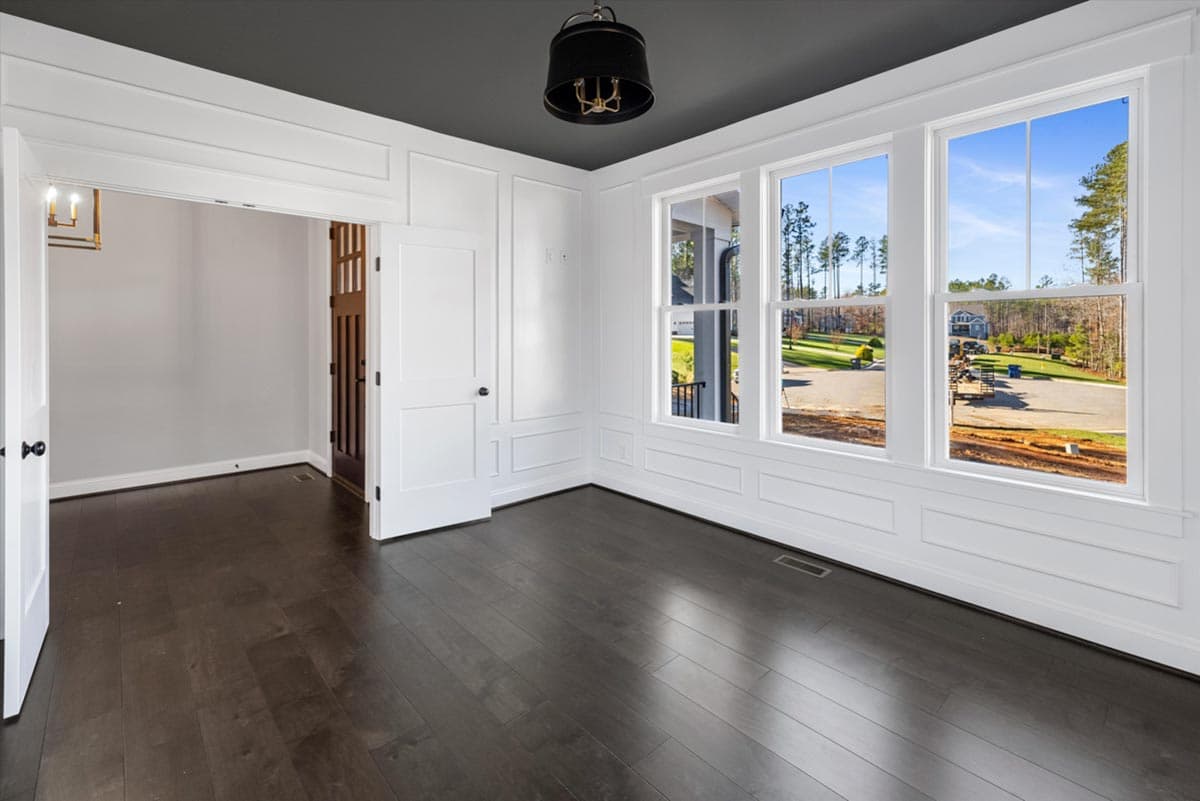 Interior view of a room with dark wood floors, white wainscoting, and three large double-hung windows overlooking a street.