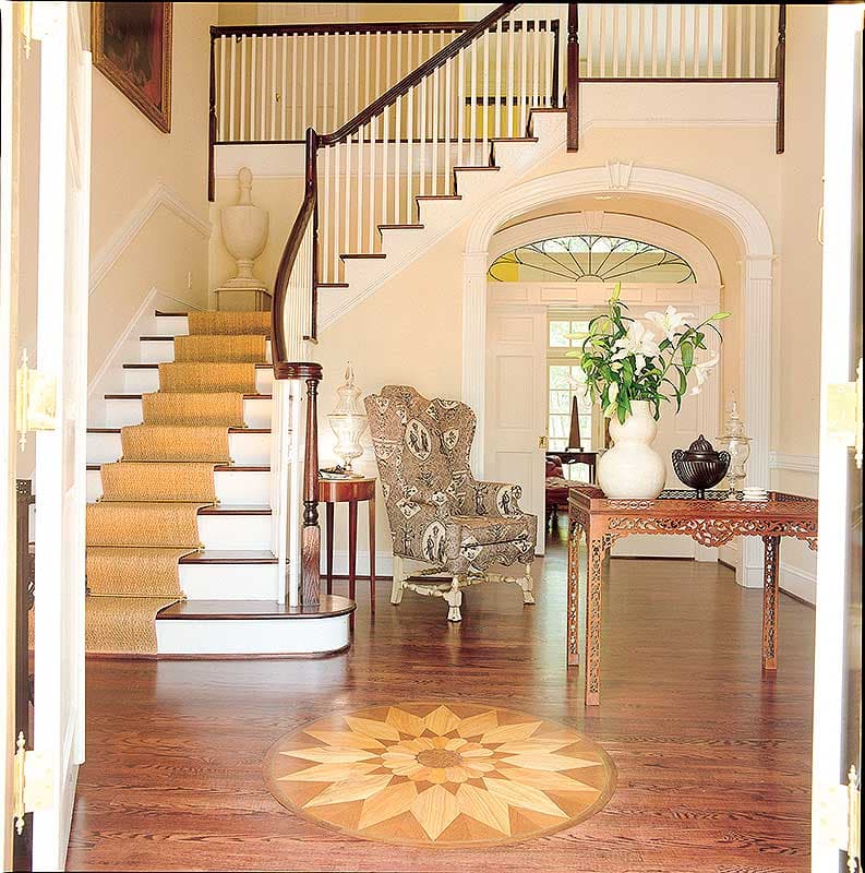 Foyer with curving staircase, patterned armchair, ornate table, and decorative wood inlay floor.