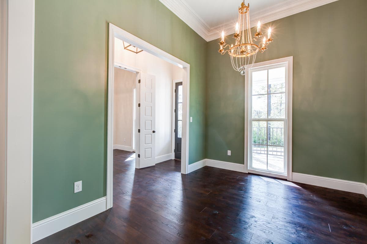 Interior view of room with green walls, chandelier, tall window, dark wood floor, and doorway to hallway.